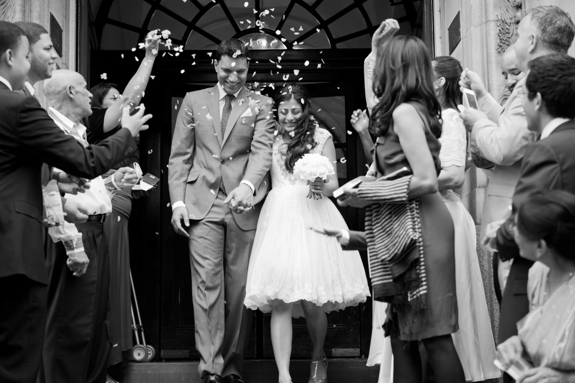 A newly married couple leaving Chelsea Town Hall surrounded by their friends and family