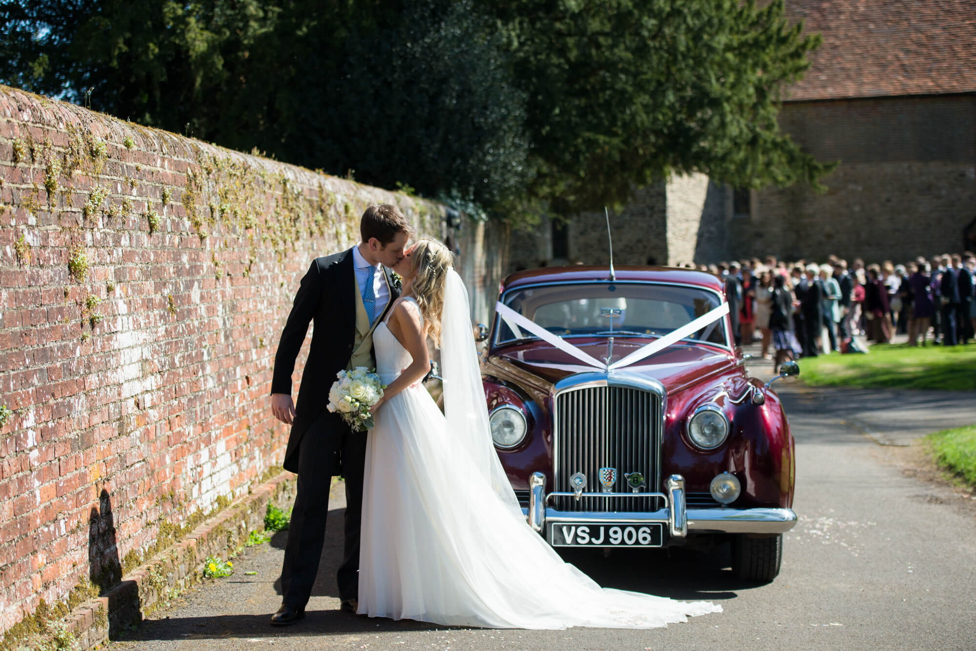 A bride kissing her husband in front of a vintage car with a ribbon tied on it